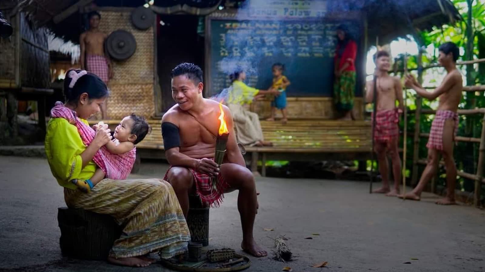 A family in traditional dress in a heritage village, with a woven house and old script signs behind