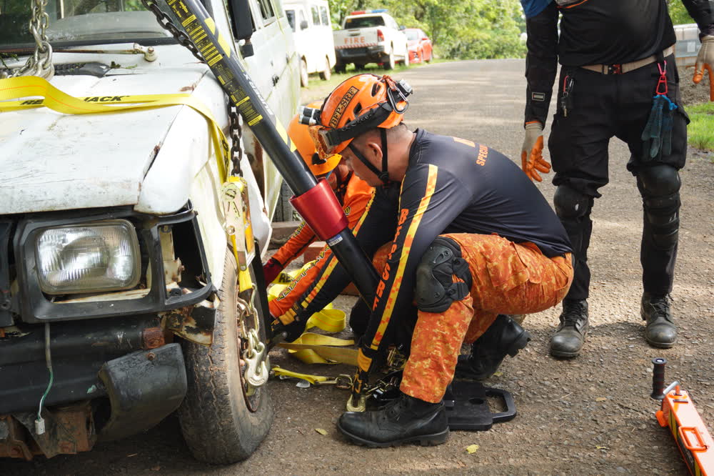 Rescuers in orange gear using hydraulic tools on a vehicle in a training drill