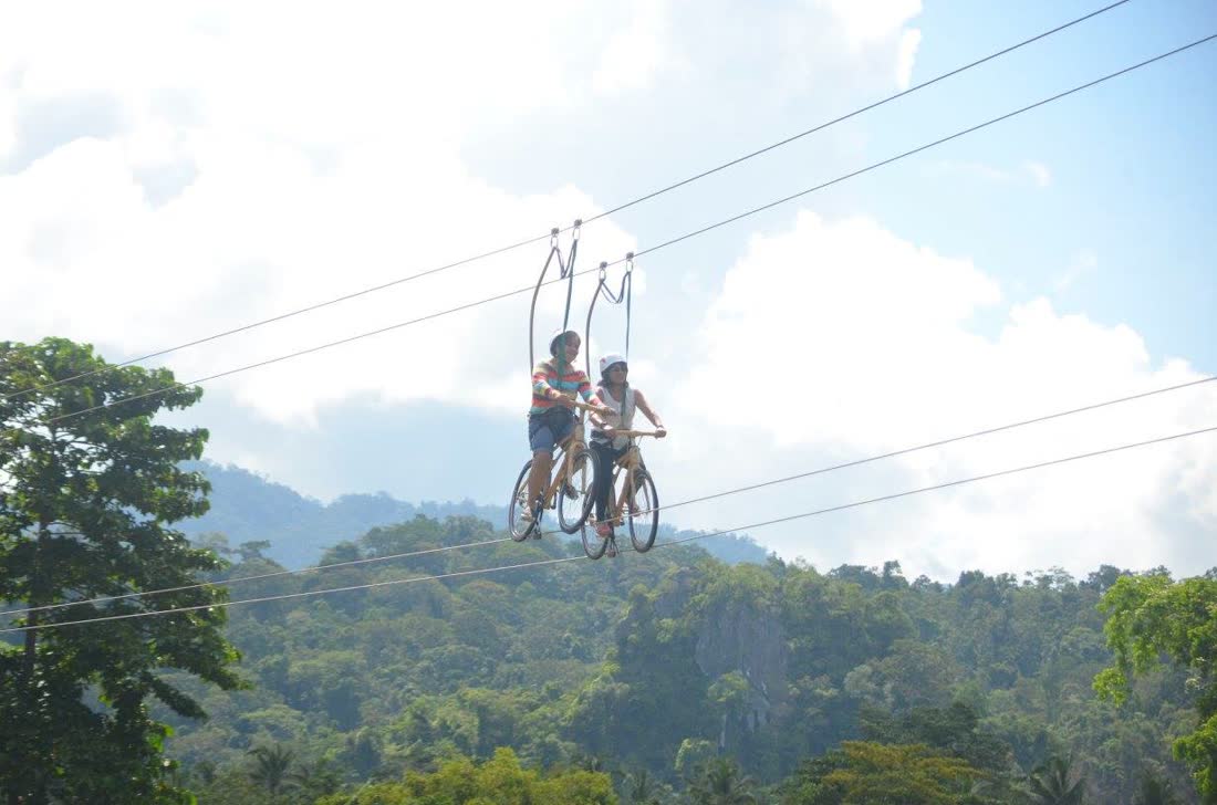 Two riders pedalling a bicycle along a cable high above the trees