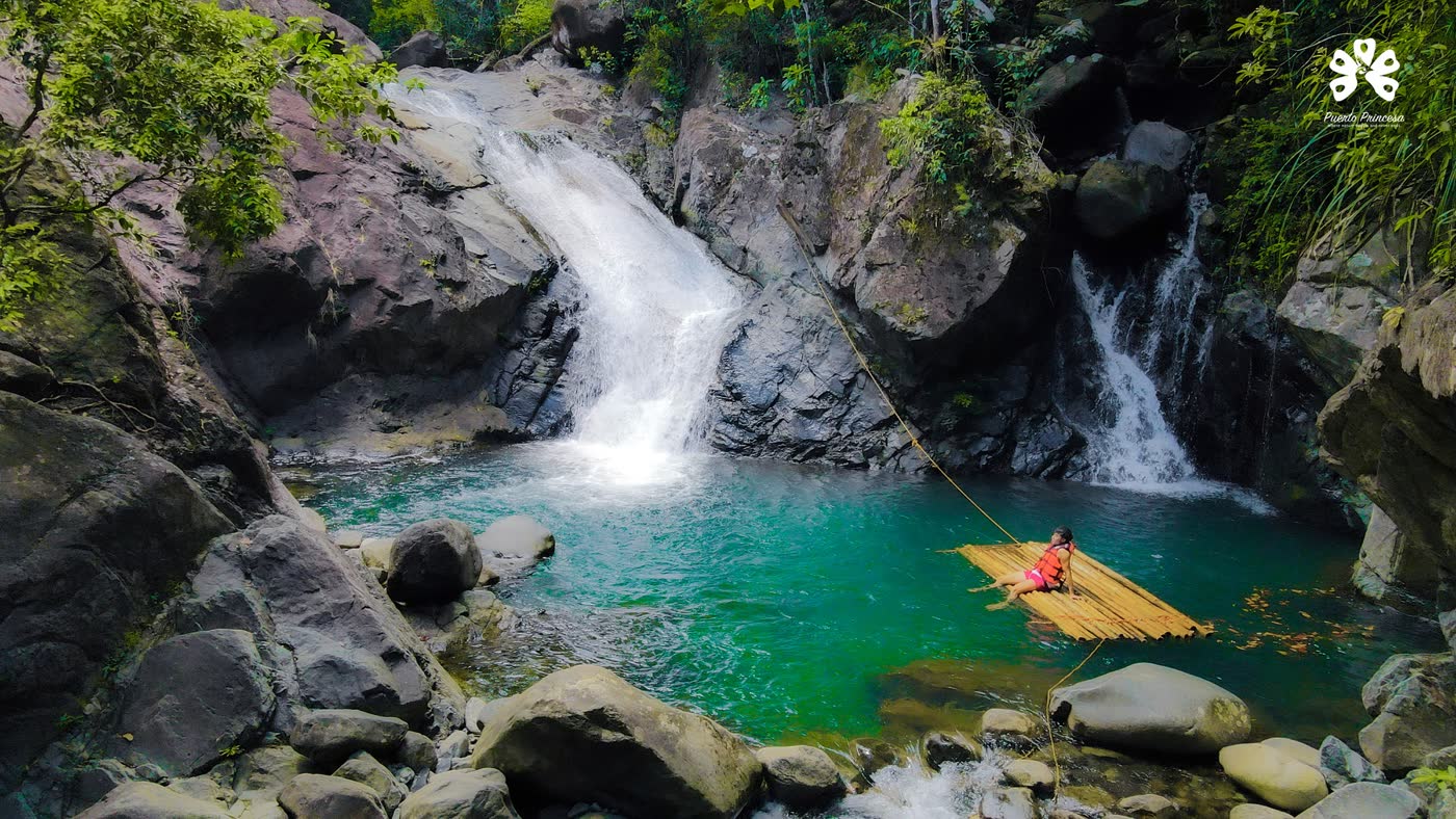 A waterfall pouring into a clear green pool between boulders, a bamboo raft on the water
