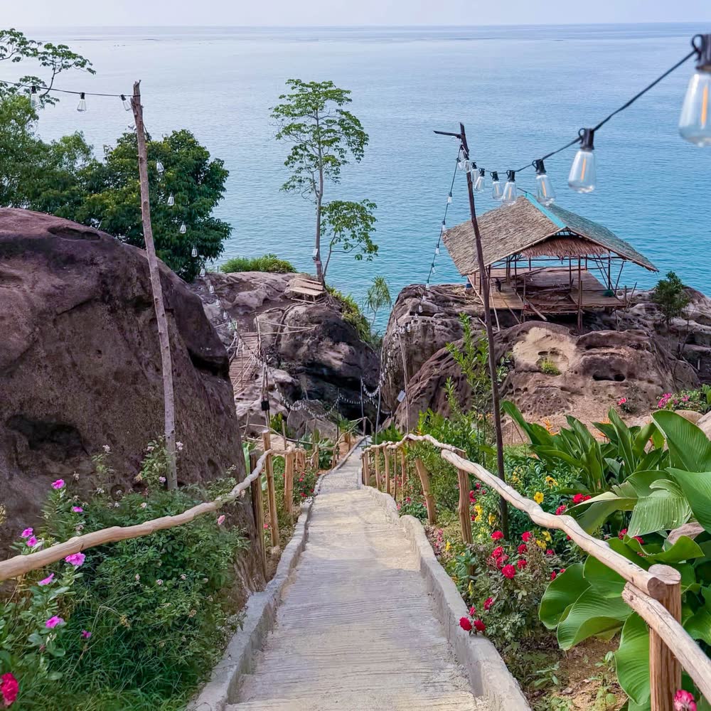 A stone path lined with flowers leading down to a thatched hut by the sea