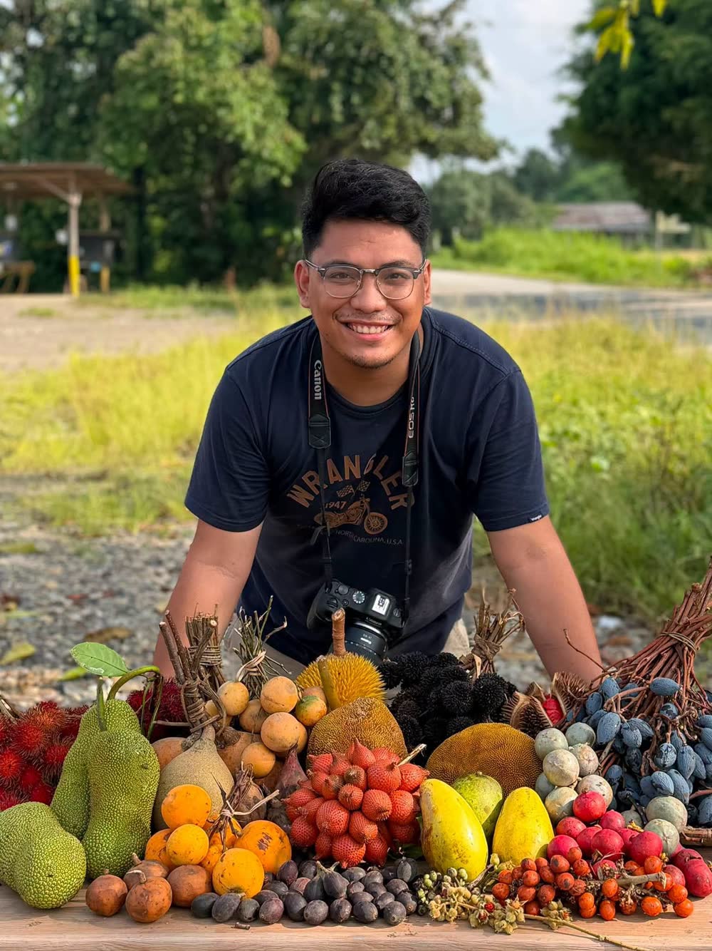 A smiling man with a camera behind a table of durian, lanzones and other fruit