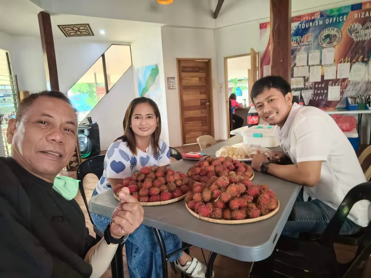 Three people at a table piled with red rambutan, smiling for the camera