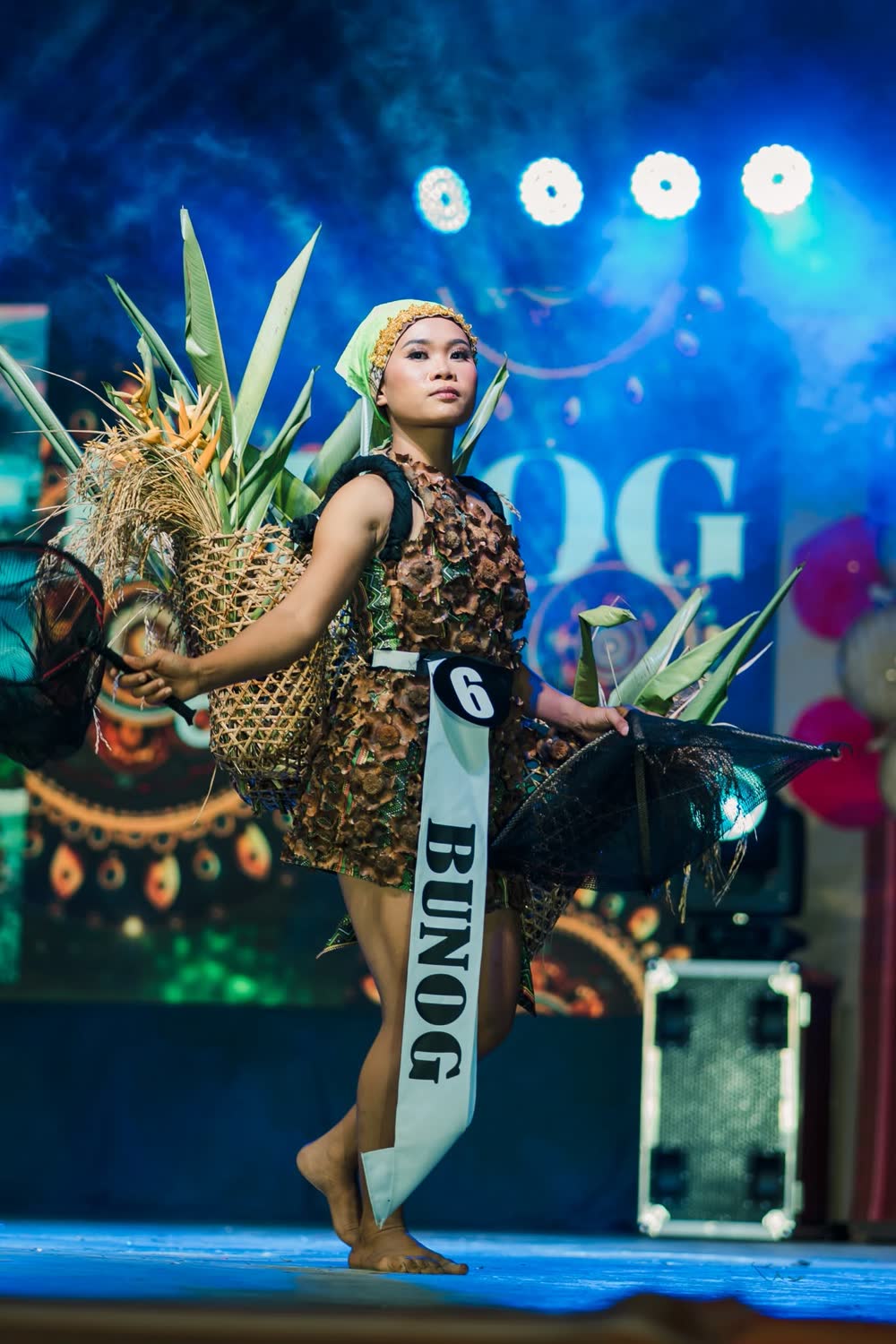A candidate with a Bunog sash in a woven costume holding a basket