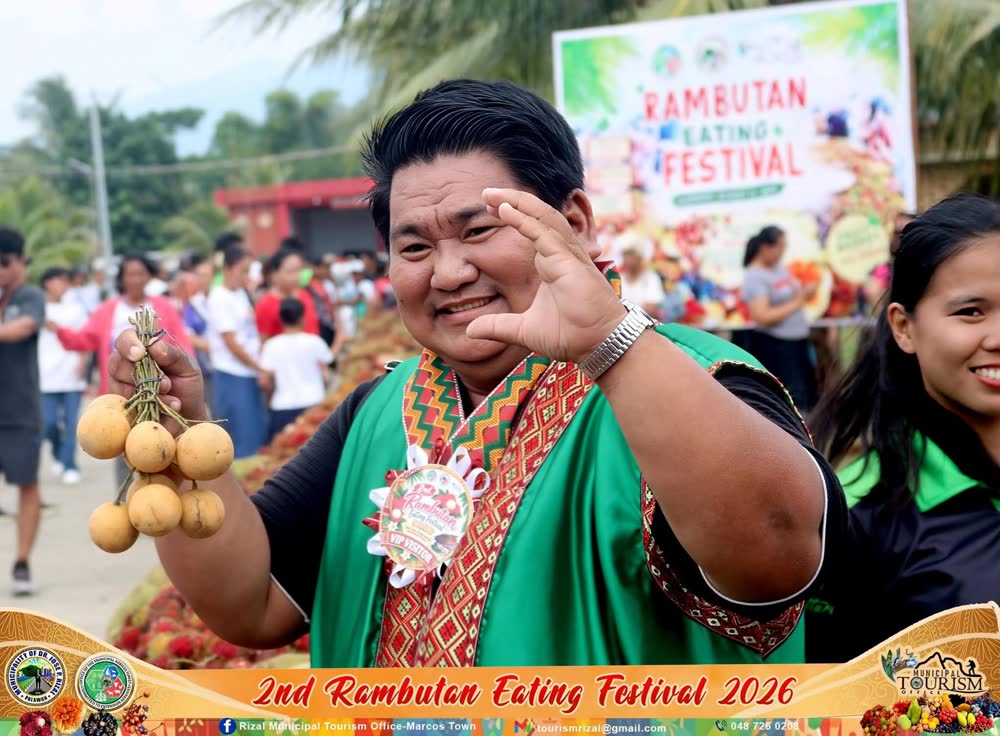 A smiling man in a green and red woven vest holds up a bunch of fruit at the Rambutan Eating Festival