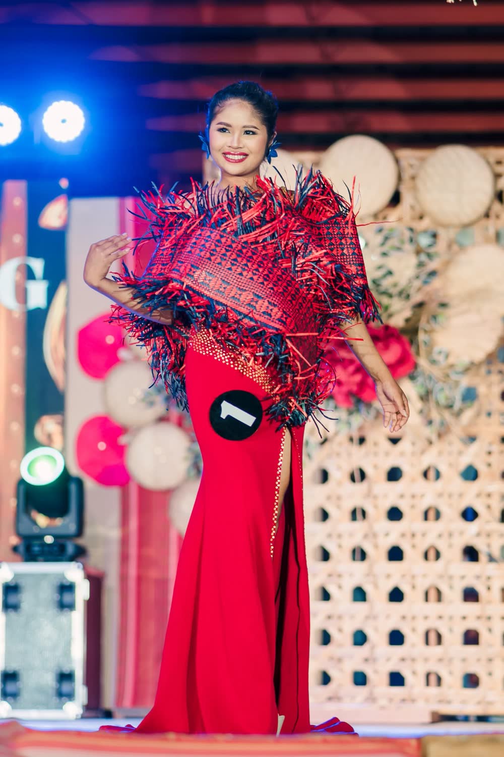 A candidate in a red gown with a woven fringed cape