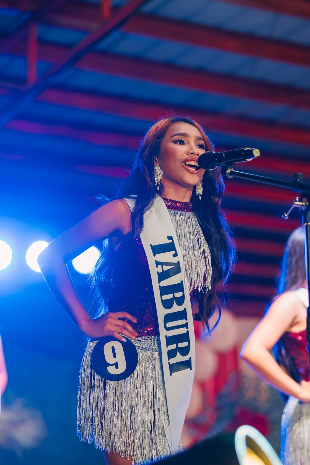 A candidate wearing a Taburi sash sings into a microphone on stage