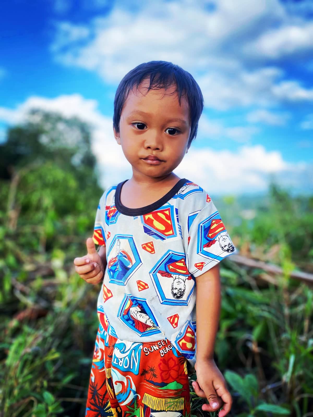 A small boy in a Superman shirt standing against a blue sky and green hills