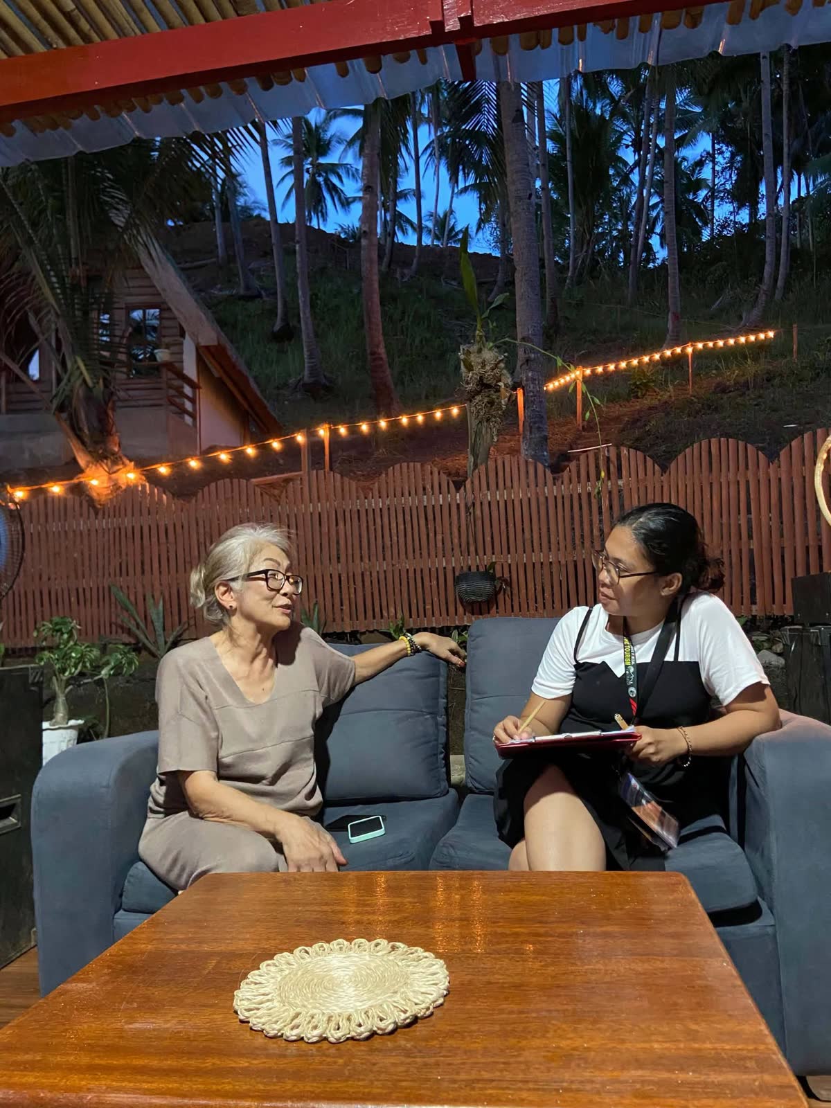 A woman taking notes while interviewing an older woman on a porch at dusk