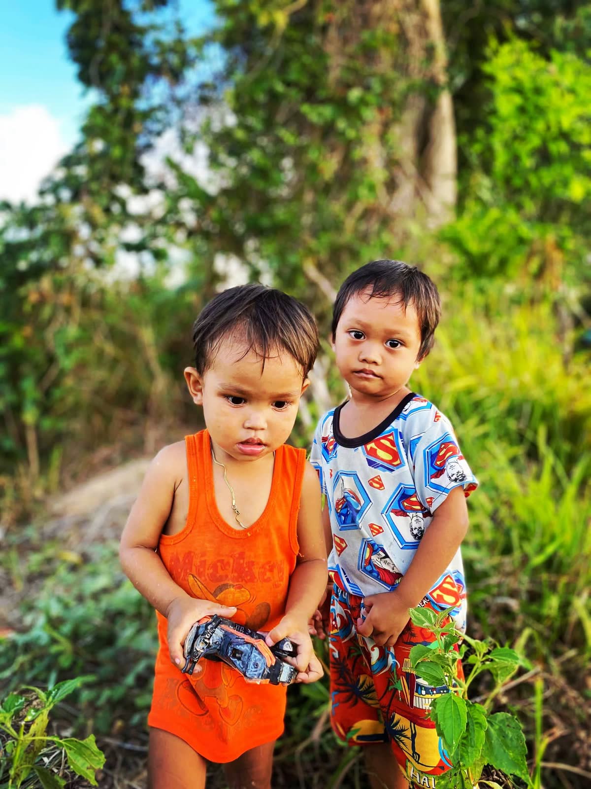 Two small children standing together among green leaves, looking at the camera