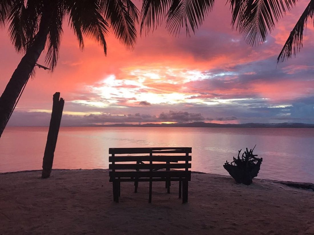 A wooden bench on the beach facing a pink sky over the sea