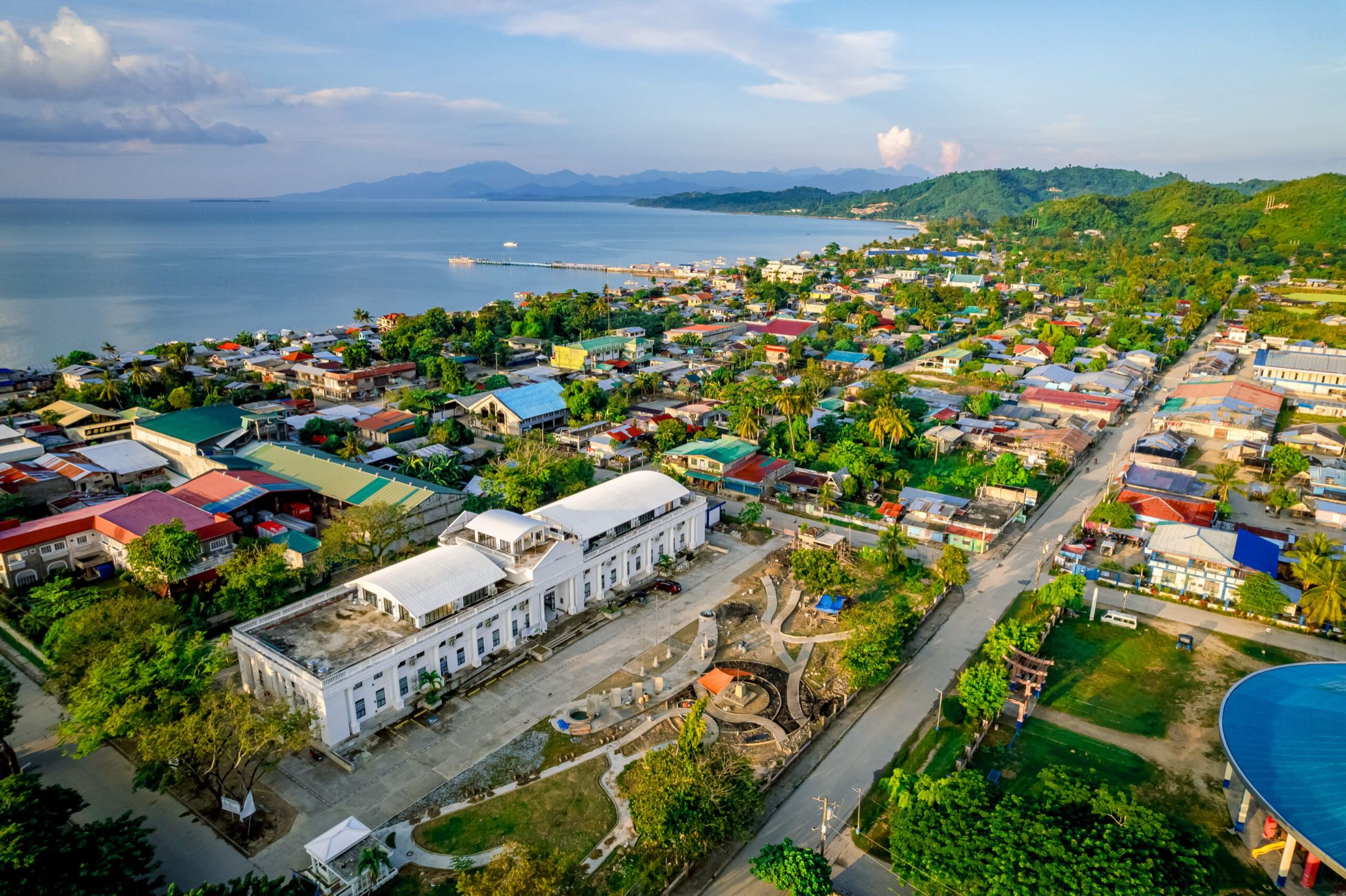 Aerial view of Roxas at golden hour: the white Municipal Hall and plaza, the town, the sea and the mountains beyond