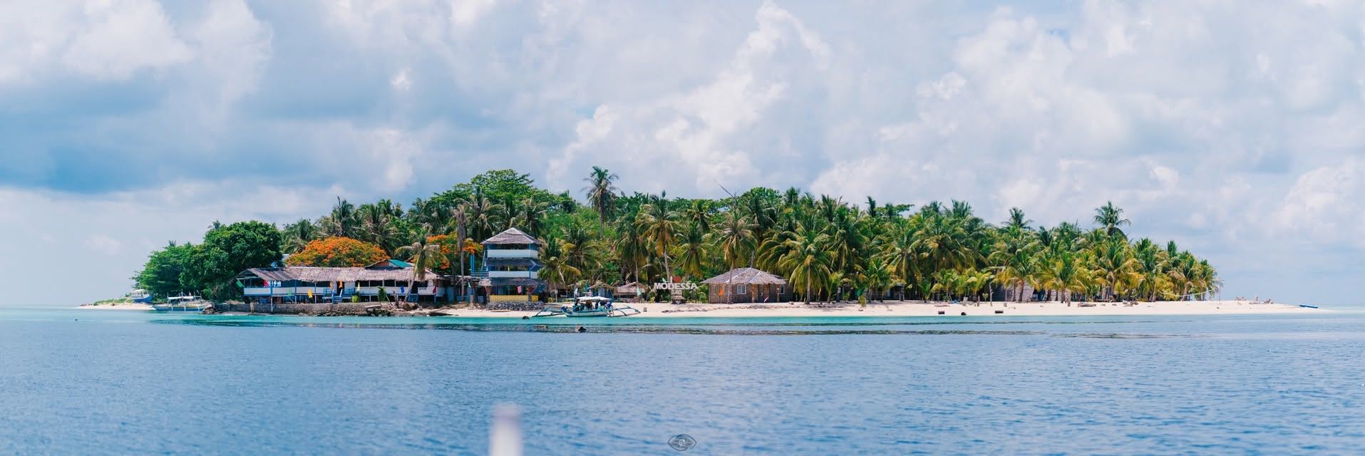 An island resort seen from the water: palms, cottages and a long jetty
